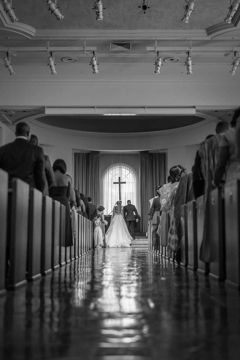 This image, artfully captured by Chris Spicks Photography, a Houston wedding photographer, depicts a poignant moment in a wedding ceremony. The scene is framed within the elegant confines of a church aisle, leading the eye to the couple at the altar. Guests line the pews on either side, their postures suggesting rapt attention to the vows being exchanged.  The photograph is rendered in timeless black and white, emphasizing the contrasts and textures from the polished floor to the intricate details of the ceiling. The grand window at the end of the aisle bathes the couple in a soft, natural light, creating a symbolic and serene backdrop.  The groom stands tall and poised, facing the bride, whose wedding gown flows behind her, its train spreading gracefully on the floor. The high ceiling of the venue with mounted lights and classical design elements adds a feeling of both grandeur and intimacy to the occasion.  As a Houston wedding photographer, Chris Spicks Photography has expertly captured not just a moment, but the essence of the couple’s special day, creating a wedding portrait that will be cherished for years to come. This image stands as a testament to their ability to encapsulate the emotion and elegance inherent in such a significant life event.