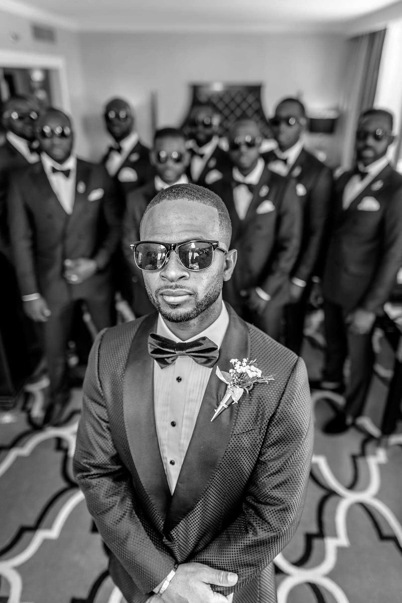 This striking black and white image, captured by Chris Spicks Photography, a renowned Houston wedding photographer, features a confident groom in the foreground, sharply dressed in a textured tuxedo with a bow tie and a classic boutonnière. His attire is elegantly complemented by sunglasses, adding a touch of modern sophistication to his wedding day ensemble.  Standing in the background, slightly out of focus to emphasize the groom, is his entourage of groomsmen. They are uniformly dressed in sleek tuxedos, mirroring the groom's attire, each sporting sunglasses as well, which lends a cool, cohesive look to the group. Their poise and the unity in their outfits create a powerful visual statement.  The composition of the photo is masterful, with the groom centered and his groomsmen fanned out behind him, creating a sense of depth and dimension in the photo. The carpet's intricate pattern adds a dynamic texture to the scene, contrasting with the smooth lines of the groomsmen's tuxedos.  Chris Spicks Photography has skillfully used the depth of field to draw focus to the groom while still capturing the bond and camaraderie of the groomsmen, showcasing their expertise as a Houston wedding photographer. This image not only highlights the celebratory atmosphere but also encapsulates the style and personality of the groom and his closest friends on this momentous occasion.