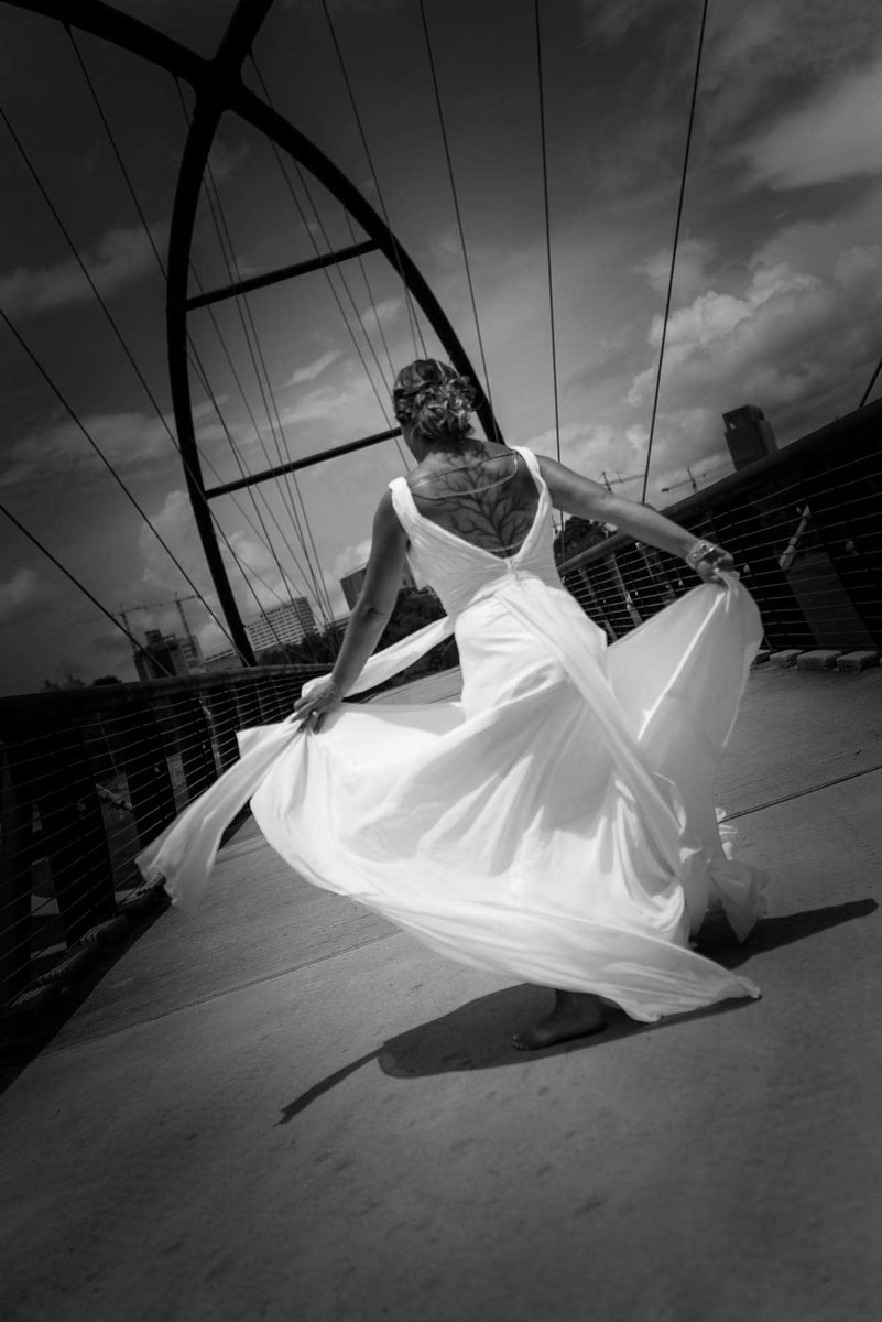In this dynamic black and white photograph by Chris Spicks Photography, a Houston wedding photographer, we see a bride captured in a moment of spontaneous joy. She is standing on a bridge, her wedding dress in motion, creating a fluid arc that mirrors the lines of the bridge's structure above her. The bride's back is to the camera, allowing us to admire the intricate details of her dress and the elegant updo of her hair, which is complemented by a delicate hairpiece.  The strong geometric lines of the bridge contrast with the soft swirl of the dress, and her pose—arms outstretched and one leg visible in the swirl of fabric—conveys both grace and excitement. The clouds in the sky above and the urban skyline hint at the city of Houston's backdrop, suggesting a narrative of a special day spent in the heart of the city.  This image is a testament to the artistry of Chris Spicks Photography, showcasing their ability to capture not just a wedding portrait but the essence of a bride's joy on her wedding day. The choice of black and white tones adds a timeless quality to the image, focusing on the interplay of light, shadow, and texture. It's a photograph that tells a story and evokes emotion, embodying the celebratory spirit of a wedding captured by a skilled wedding photographer in Houston.