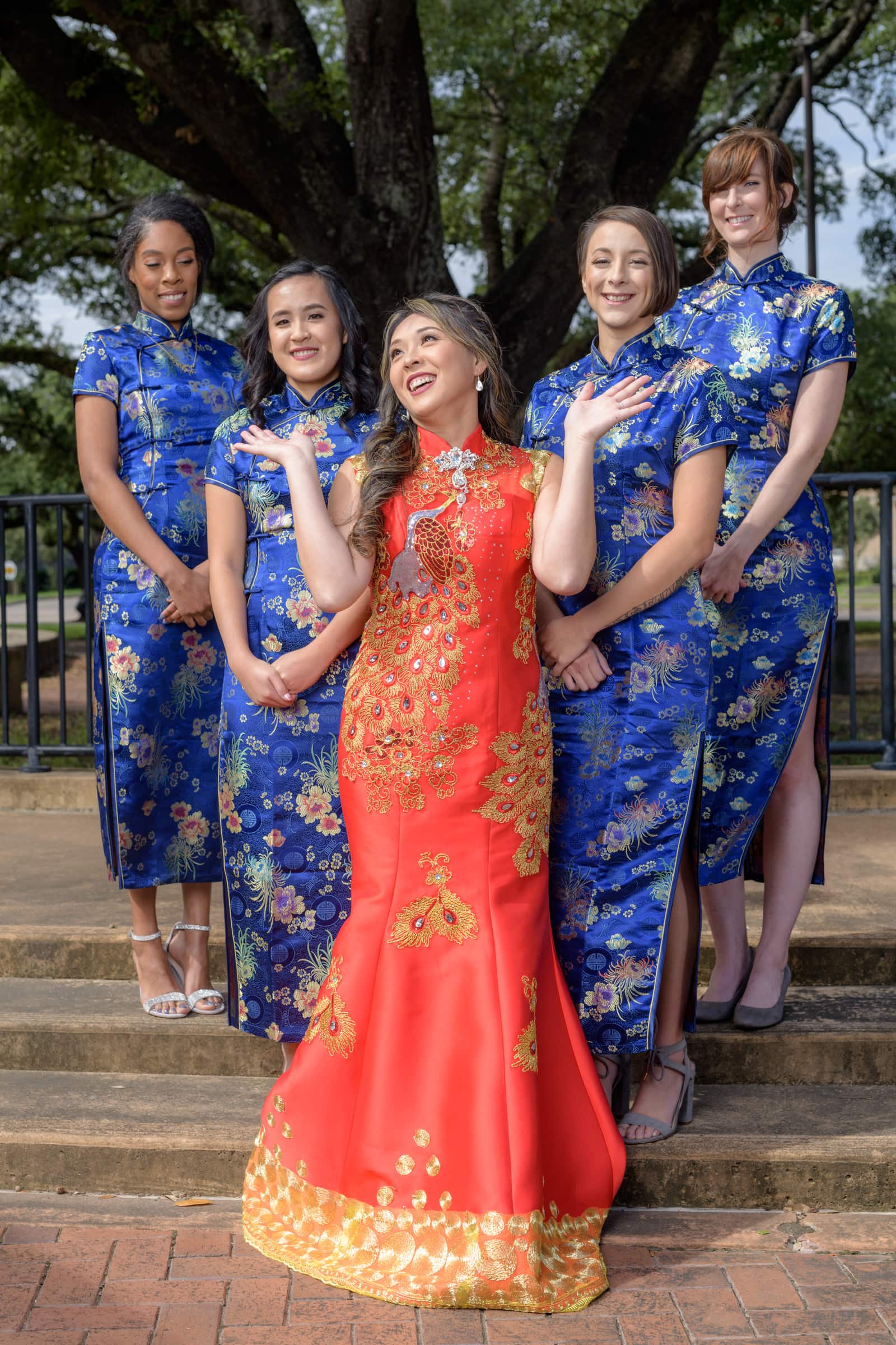  In this vibrant and colorful photograph taken by Chris Spicks Photography, known for their exceptional Houston wedding photographer services, we are presented with a beautiful moment that blends cultural tradition with the joy of a wedding celebration.  The image features a bride at the center, radiant in a traditional red gown adorned with intricate golden embroidery, which symbolizes good luck and happiness in many Eastern cultures. Her attire is stunning, with a high collar and fitted bodice that flows into a full skirt, culminating in a golden hem. The bride's hair is styled elegantly, and she wears a smile that lights up the frame, expressing her excitement and joy on this special day.  Flanking the bride are her bridesmaids, each dressed in harmonious blue cheongsam dresses with floral patterns, their smiles reflecting the festive spirit. The unity in their attire creates a beautiful contrast against the bride’s red gown, adding depth and richness to the visual composition.  The backdrop of a large, lush tree and the clear skies above, as captured in this outdoor setting, adds a natural serenity to the scene. The light cascades softly over the bridal party, illuminating them with a gentle glow that highlights the colors and textures of their exquisite garments.  This photograph is a testament to the skill and artistry of Chris Spicks Photography, showcasing their ability to capture not just the visual splendor of a wedding but the cultural essence and personal happiness of the bride and her close companions. It’s an image that tells a story of tradition, friendship, and the start of a new chapter, all within the heart of Houston.