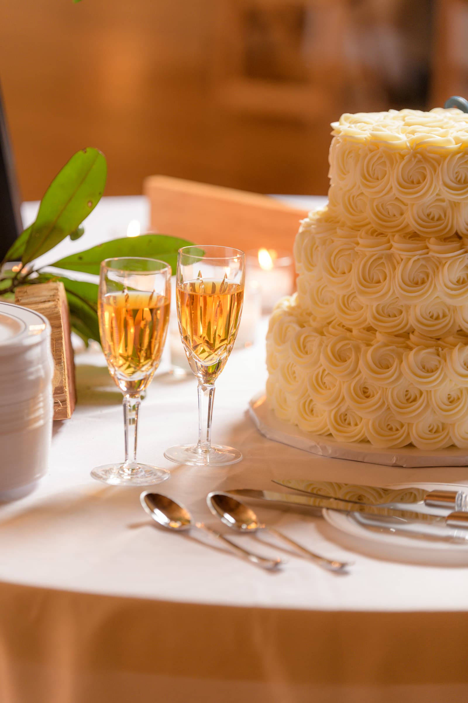 This exquisite photograph, captured by the expert team at Chris Spicks Photography, a distinguished Houston wedding photographer, features a beautifully composed still life from a wedding reception. The warm glow of the image highlights a table set for celebration, with two flutes of sparkling champagne sitting elegantly in the foreground. Their golden hues are rich and inviting, reflecting the joyous ambiance of the occasion.  Behind the champagne glasses, the wedding cake stands as a centerpiece, adorned with intricate swirls of creamy frosting that spiral delicately around its tiers. The craftsmanship of the cake's design is impeccable, displaying the kind of attention to detail that Chris Spicks Photography is known for capturing in their work.  The placement of the objects, from the silverware to the glasses and the natural greenery accent beside the cake, is meticulous, creating a picturesque tableau that beckons guests to partake in the festivities. The soft, natural lighting enhances the textures and colors, imbuing the scene with a sense of warmth and intimacy.  As a Houston wedding photographer, Chris Spicks Photography has a keen eye for such details, expertly preserving the beauty and elegance of every aspect of a wedding day. This photograph not only captures the luxurious elements of the celebration but also the ambiance of sophistication and love that surrounds the couple's special day in Houston.