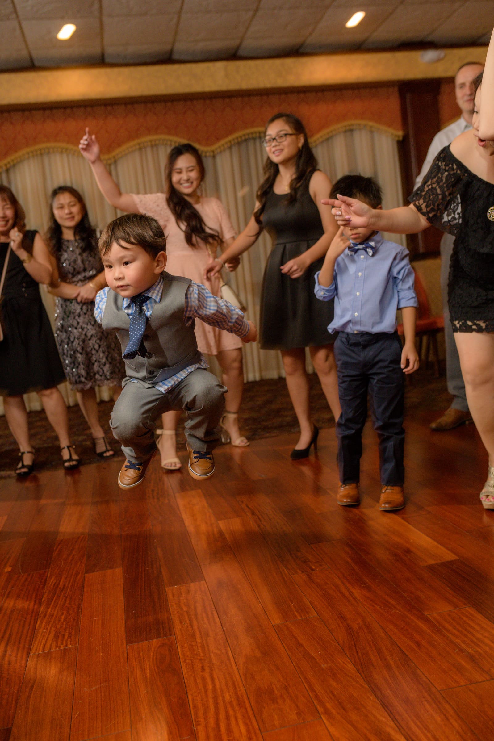 Captured by the talented team at Chris Spicks Photography, a Houston wedding photographer, this image is vibrant with the joy and energy of a wedding celebration. It features a lively moment on the dance floor with young children fully immersed in the festivities.  In the center of the frame, a young boy in mid-jump steals the spotlight. He's sharply dressed in a blue shirt with a matching vest and tie, complete with rolled-up sleeves and stylish shoes, exuding the carefree spirit and excitement of the occasion. His expression is one of pure concentration and delight as he dances.  Beside him stands another boy, slightly older, dressed in a smart blue shirt and navy pants. He appears more reserved, perhaps momentarily overwhelmed by the bustle around him, shielding his eyes from the flash of the camera or the lights of the dance floor.  The background is filled with guests of varying ages, all participating in the dance. Their blurred movements suggest the rhythm and pace of the music, adding to the dynamic atmosphere of the scene. The warm tones of the wooden floor and the soft, ambient lighting contribute to the inviting ambiance.  This photo exemplifies Chris Spicks Photography's ability to capture candid, heartfelt moments at Houston weddings. It's not just the formal portraits that tell the story of a wedding day, but also these spontaneous bursts of joy, especially from the youngest attendees, that add character and charm to the celebration's narrative.