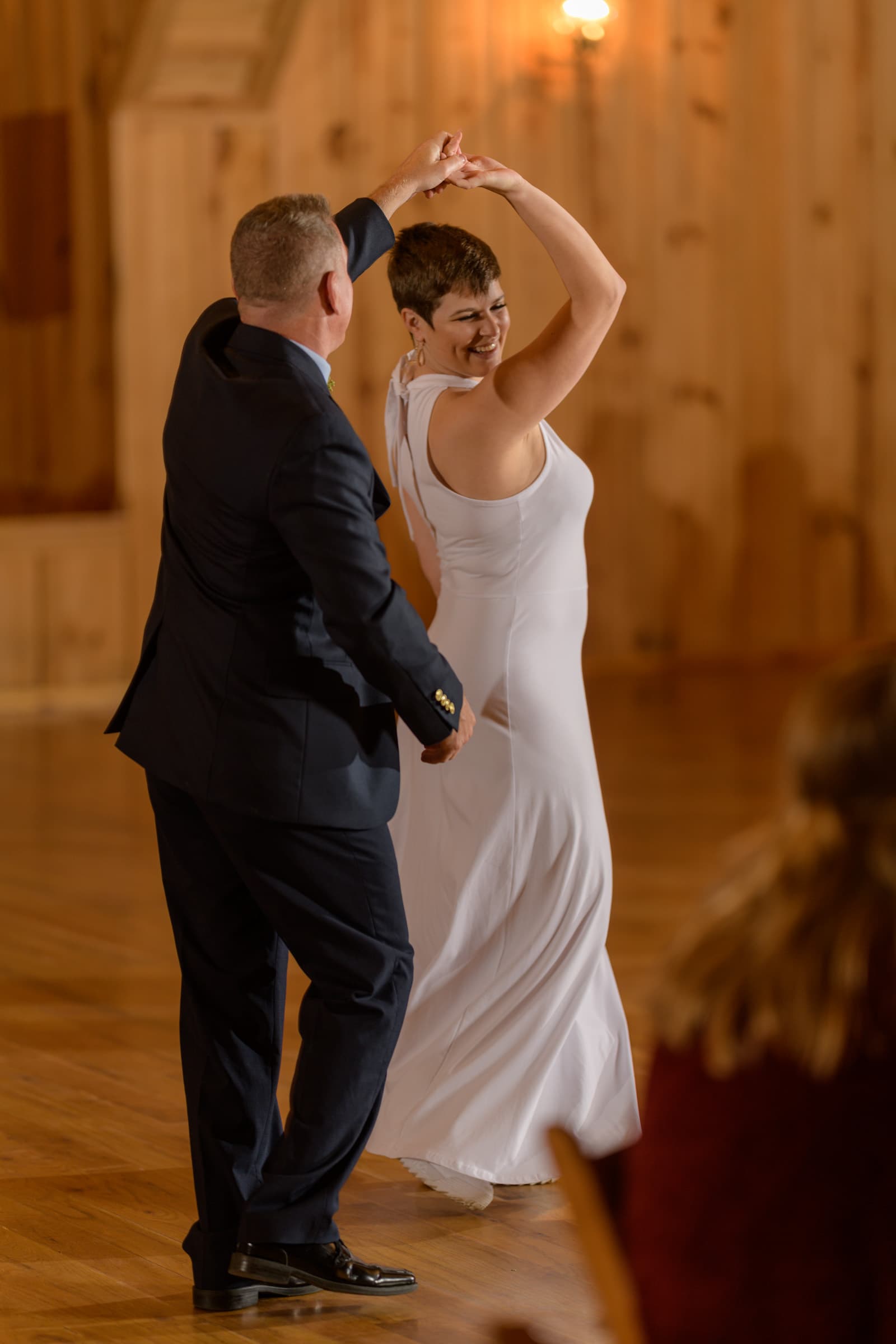  This heartwarming photograph by Chris Spicks Photography, renowned for their Houston wedding photographer expertise, captures a timeless moment of joy and affection between a father and daughter during a wedding dance. The warmth of the wooden interior, likely a rustic venue in Houston, sets a cozy backdrop for this tender scene.  In the image, the father, smartly dressed in a classic dark suit, leads his daughter in a dance. His expression is one of pride and happiness as he spins his daughter, whose wedding dress swirls around her. The bride, sporting a chic, short haircut and a radiant smile, looks up at her father with a look of love and gratitude. Her elegant, sleeveless white gown and the fluid motion of the fabric accentuate the movement and emotion of the dance.  The soft lighting from above enhances the intimate atmosphere, and the candid shot captures the genuine connection and joy shared by the family on this significant day. The photograph not only preserves a significant moment but also reflects the skill of Chris Spicks Photography in encapsulating the essence of a wedding day's celebration, showcasing their ability to create lasting memories for families in Houston and beyond.