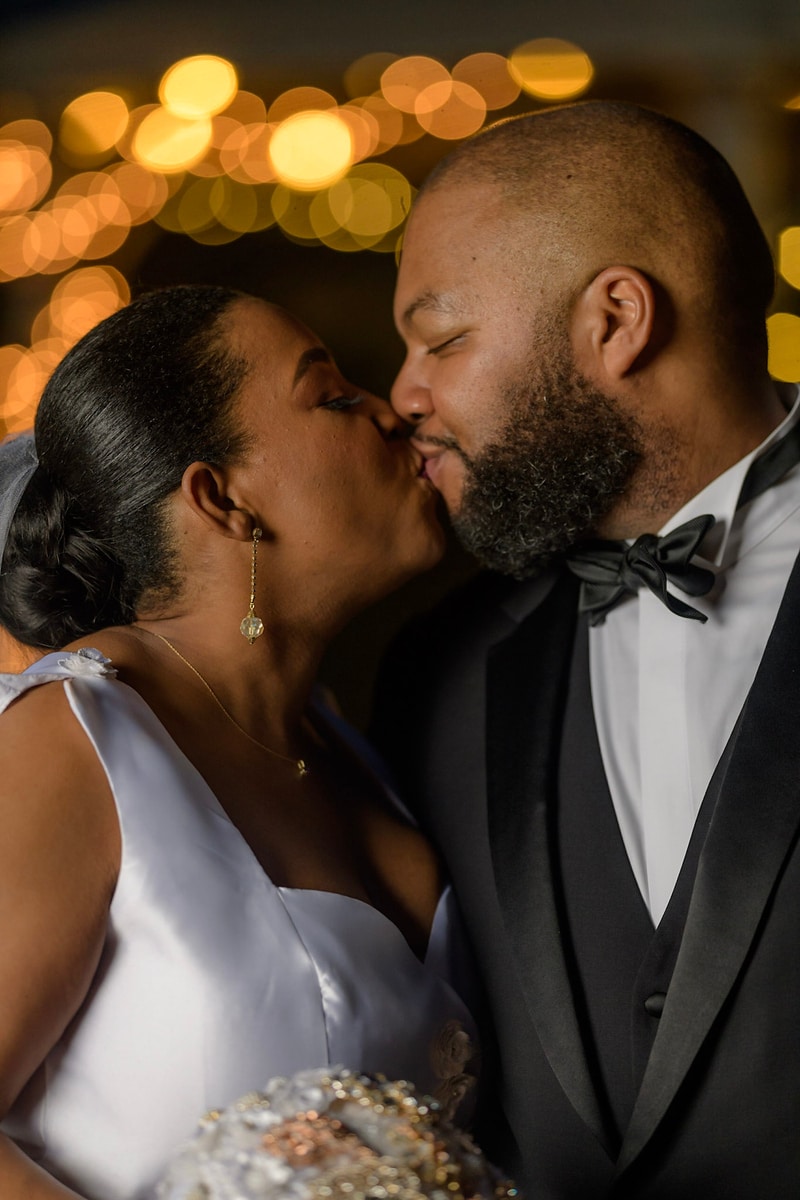 This image, expertly captured by Chris Spicks Photography, showcases the intimate moment of a couple sharing a kiss on their wedding day. As a premier Houston wedding photographer, they have focused on the couple's connection, bringing to life the love and tenderness shared between the two.  The photograph features the bride and groom in a close embrace, their lips meeting in a gentle kiss. The bride's earrings dangle elegantly, catching the light as she leans in for the kiss, and her gown is detailed with delicate beading that adds a touch of glamour to the moment. The groom, in a sharp tuxedo with a black bow tie, reciprocates with equal affection, his beard neatly groomed for their special day.  Behind them, a bokeh of warm, golden lights creates a magical and romantic backdrop, symbolizing the celebratory and joyful spirit of the occasion. The lights shimmer out of focus, drawing all attention to the couple's loving gesture, a technique that showcases Chris Spicks Photography's ability to capture the essence of a wedding's magical moments.  This image not only captures the beauty and romance of the couple's special day in Houston but also the artistry that Chris Spicks Photography brings to their work, creating memorable images that their clients will treasure for a lifetime.