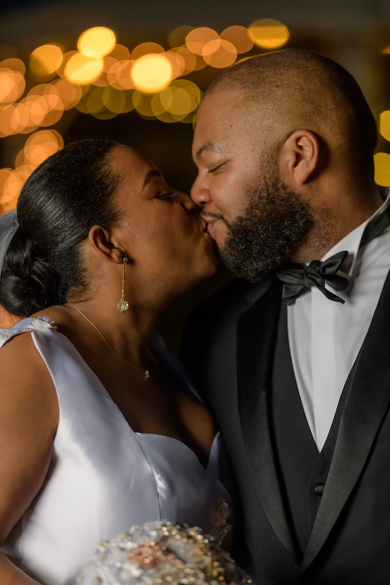 This image, expertly captured by Chris Spicks Photography, showcases the intimate moment of a couple sharing a kiss on their wedding day. As a premier Houston wedding photographer, they have focused on the couple's connection, bringing to life the love and tenderness shared between the two. The photograph features the bride and groom in a close embrace, their lips meeting in a gentle kiss. The bride's earrings dangle elegantly, catching the light as she leans in for the kiss, and her gown is detailed with delicate beading that adds a touch of glamour to the moment. The groom, in a sharp tuxedo with a black bow tie, reciprocates with equal affection, his beard neatly groomed for their special day. Behind them, a bokeh of warm, golden lights creates a magical and romantic backdrop, symbolizing the celebratory and joyful spirit of the occasion. The lights shimmer out of focus, drawing all attention to the couple's loving gesture, a technique that showcases Chris Spicks Photography's ability to capture the essence of a wedding's magical moments. This image not only captures the beauty and romance of the couple's special day in Houston but also the artistry that Chris Spicks Photography brings to their work, creating memorable images that their clients will treasure for a lifetime.