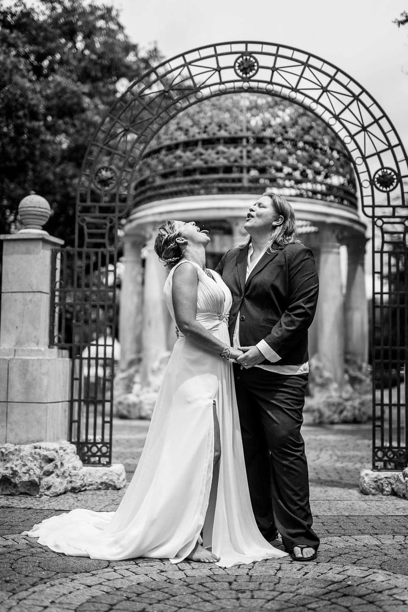 This emotive black and white image, expertly captured by Chris Spicks Photography, a Houston wedding photographer, showcases a couple on their wedding day, sharing a moment of pure joy and laughter. The photograph is set against an elegant wrought iron gazebo, adorned with intricate patterns that add a classic touch to the composition.  The brides, standing close together on a cobblestone path, are lost in a moment of shared mirth. One bride, dressed in a sleek white gown with a graceful train, tilts her head back capturing raindrops on her tongue, her hand clasped in her partner's. Her spouse, wearing a smart black suit paired with an open-necked white shirt, looks down at her with an expression of adoration and shared happiness.  The light in the photograph highlights the brides' expressions and the details of their wedding attire, from the gentle drape of the gown to the relaxed elegance of the suit. The joyous atmosphere is palpable, as the surrounding architecture and the open sky above frame this intimate scene.  As a Houston wedding photographer, Chris Spicks Photography has a gift for capturing the unguarded, spontaneous moments that truly reflect the spirit of a couple's special day. This image is a celebration of love, capturing the beauty of the moment and the essence of the couple's bond in a timeless and evocative way. It's a testament to the inclusive vision that Chris Spicks Photography brings to their craft, celebrating the love of all couples in Houston and beyond.