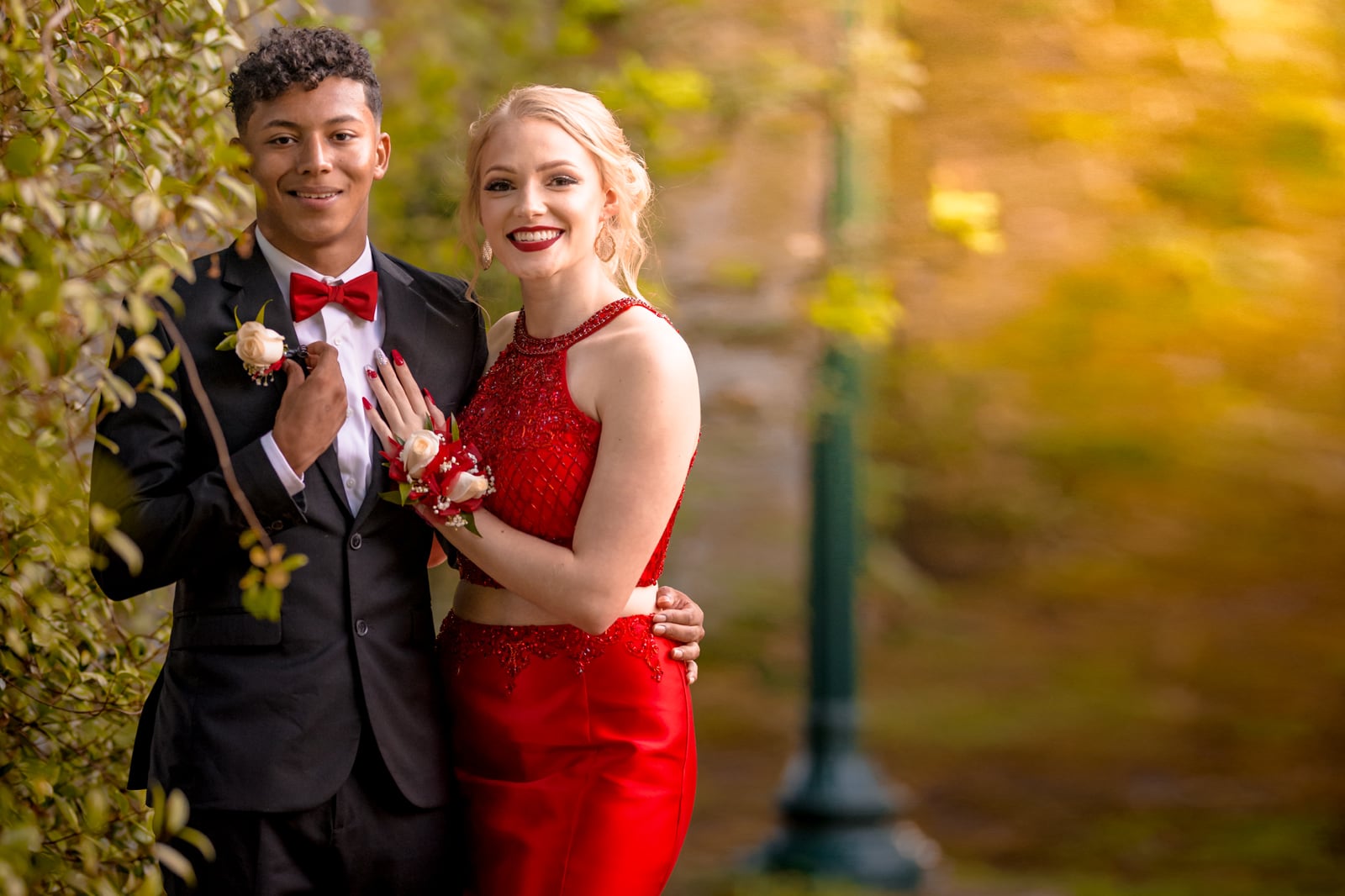 prom dates in suit and red dress pose next to green plant fence with light post in background