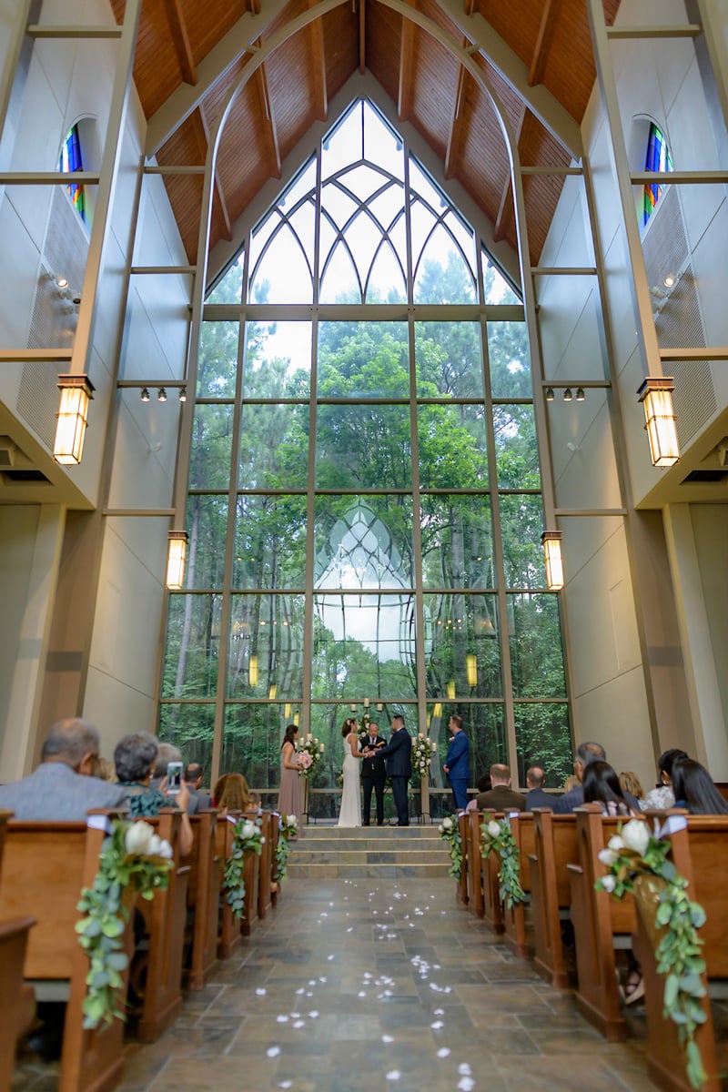 This image, captured by the talented team at Chris Spicks Photography, showcases a serene and luminous wedding ceremony within a beautifully designed chapel in Houston. As a Houston wedding photographer, the ability to encapsulate such an atmosphere is crucial, and this photo does so masterfully.  The chapel's architecture draws the eye, featuring a soaring glass window that frames the verdant greenery outside, creating a stunning natural backdrop for the couple's special moment. The high vaulted ceilings with wooden beams add a sense of grandeur and openness to the space.  In the foreground, wooden pews line both sides of the aisle, adorned with elegant floral arrangements, leading up to the altar where the wedding ceremony is taking place. The aisle itself is sprinkled with flower petals, adding to the romantic ambiance.  At the altar, the couple stands hand-in-hand, exchanging vows in the presence of an officiant. They are flanked by attendants, with bridesmaids dressed in soft pink gowns, holding delicate bouquets that complement the natural tones of the setting. The guests are seated, watching the ceremony unfold, some turning their heads to view the photographer capturing the moment.  This image not only represents the skill of a Houston wedding photographer but also reflects the ethos of Chris Spicks Photography – a commitment to capturing the essence of each couple’s love story in a way that is both enchanting and authentic. This photo stands as a testament to the company's expertise in wedding portraiture, capturing the beauty and emotion of wedding ceremonies in Houston and beyond.