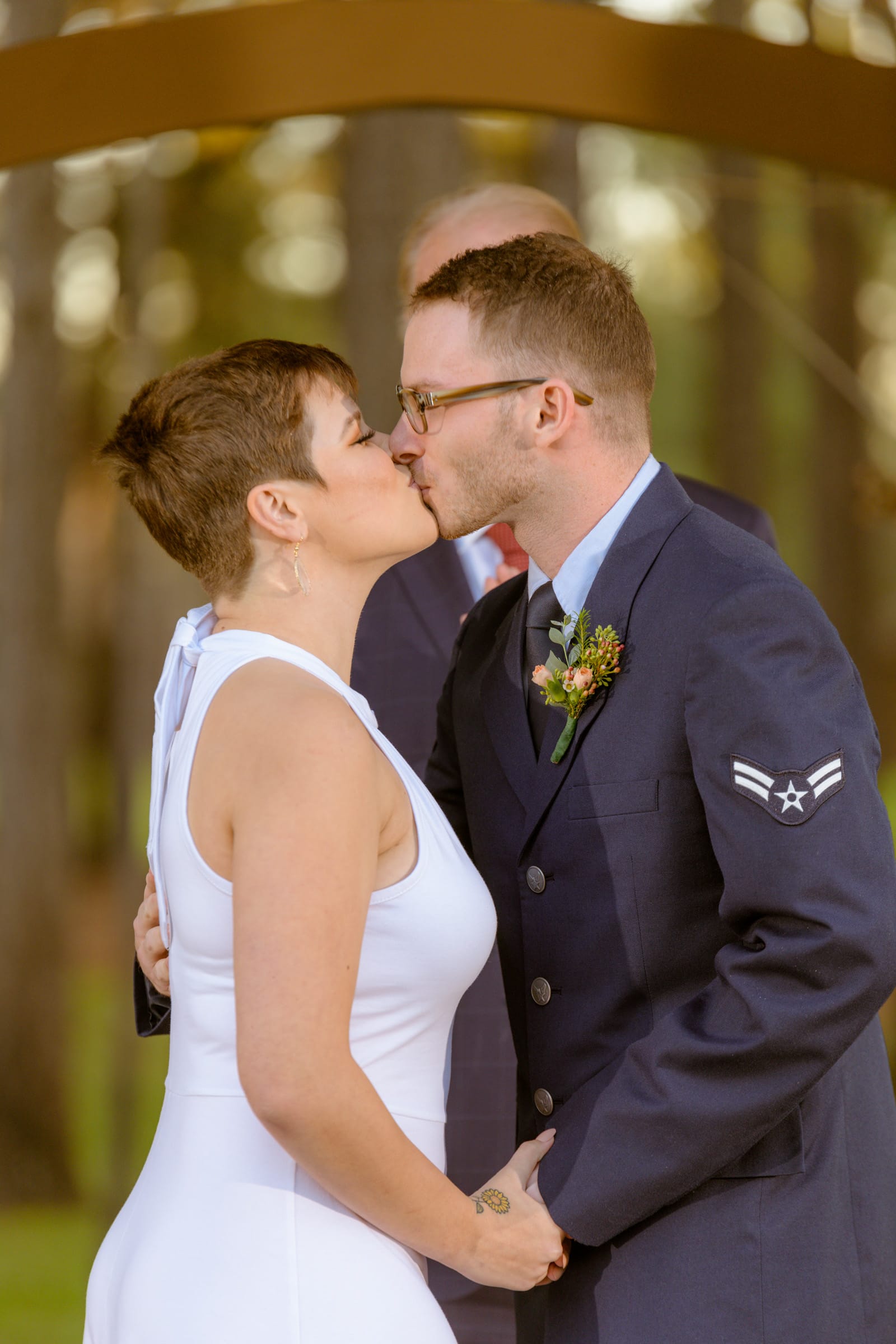 The image captures an intimate moment between a couple on their wedding day, taken by a skilled Houston wedding photographer. The setting appears to be outdoors with soft natural lighting, possibly in a park or garden, suggesting the photographer's ability to utilize Houston's diverse landscapes. The focal point is the couple engaged in a gentle kiss. On the left is a person in a sleeveless white wedding dress with a modern cut, featuring a clean, form-fitting silhouette that flatters their figure. Their hair is styled in a short, chic cut, and they wear subtle, elegant earrings. Their eyes are closed, savoring the moment of the kiss. On the right is a person in a dark military uniform, indicative of Air Force service, denoted by the chevron rank insignia on their sleeve. They also wear glasses and have a short haircut, with a focused expression and eyes closed during the kiss. A boutonnière with a mix of small flowers adds a touch of color to the dark uniform. The couple is standing closely, with the person in the dress holding the uniformed partner's hand, which gently rests on their waist. The background is a soft bokeh of greens and browns, typical of a lush, outdoor setting, creating a dreamy and romantic atmosphere. This image exudes a sense of timeless elegance and heartfelt emotion, showcasing the photographer's talent for capturing profound and tender moments that embody the spirit of a wedding day in Houston