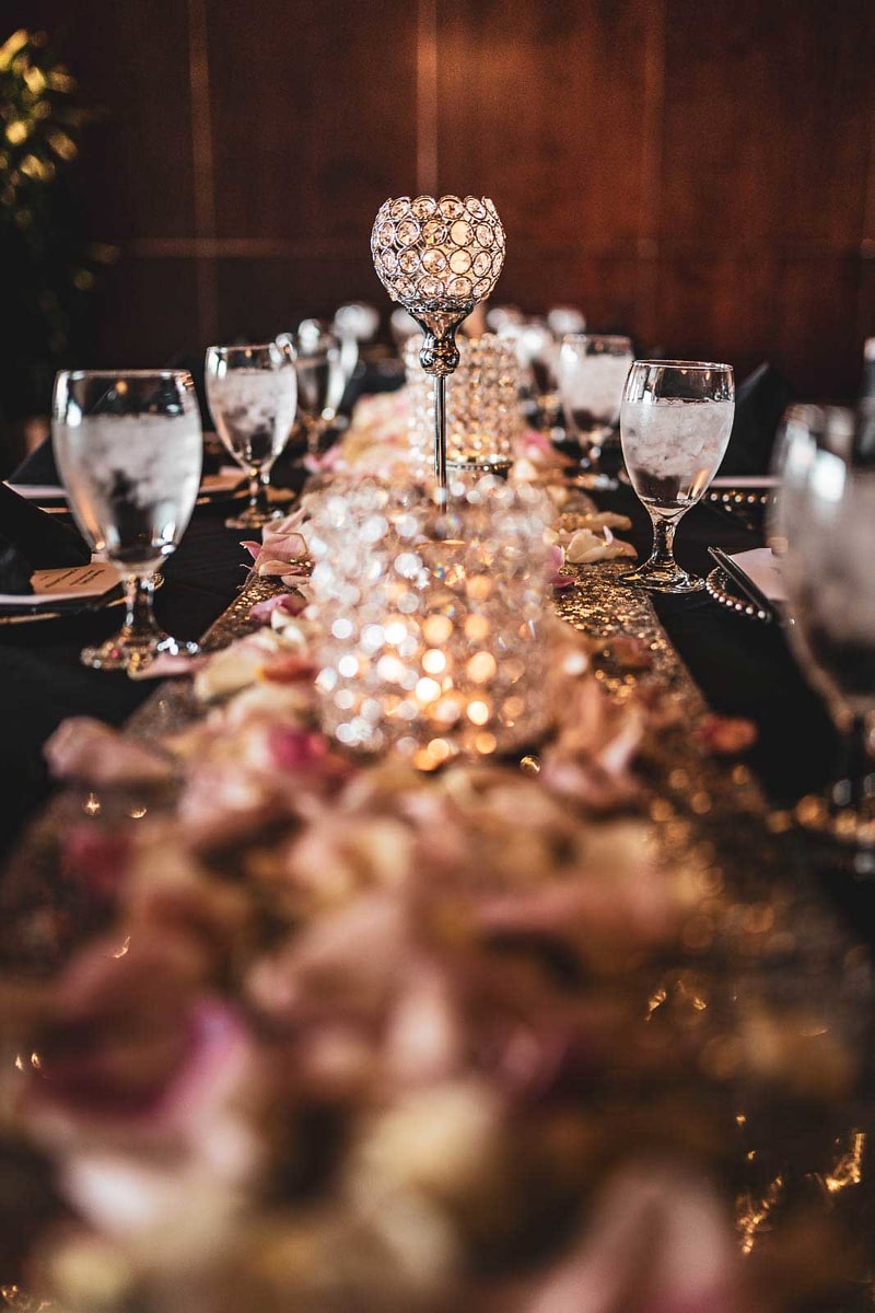 This image, taken by the skilled Houston wedding photographer at Chris Spicks Photography, beautifully captures the elegance and ambiance of a wedding reception. The table is adorned with a luxurious golden table runner that glistens under the warm light, creating a romantic and opulent atmosphere.  At the center of the image is a decorative crystal candle holder, its intricate design reflecting the surrounding lights to cast a shimmering glow across the table. This centerpiece is an exquisite focal point, surrounded by a cascade of soft rose petals and delicate floral arrangements that stretch down the length of the table.  The table setting is impeccable, with clear glasses that sparkle cleanly against the warm tones of the table, and the silverware is neatly arranged, ready for the guests to enjoy the celebration feast. Each place setting is a promise of hospitality and joy, waiting to be filled with laughter and conversation as the evening unfolds.  Chris Spicks Photography has masterfully captured not just the details of the wedding reception decor but also the mood and sophistication of a well-curated wedding event in Houston. The focus on the textures and the play of light in this image exemplifies their ability to preserve the beauty and essence of every aspect of a couple's special day.