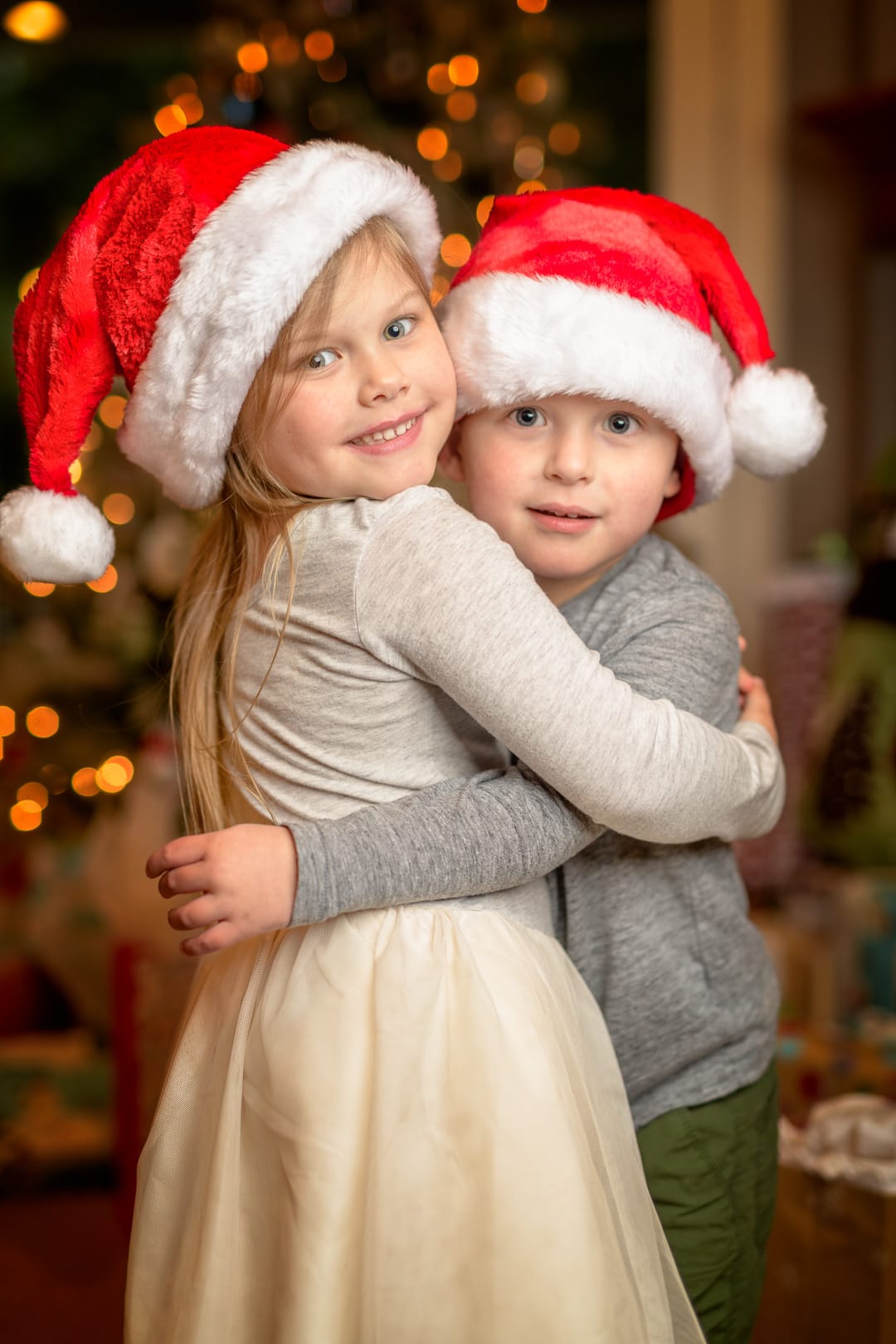 Two children in santa hats hugging in front of a christmas tree.