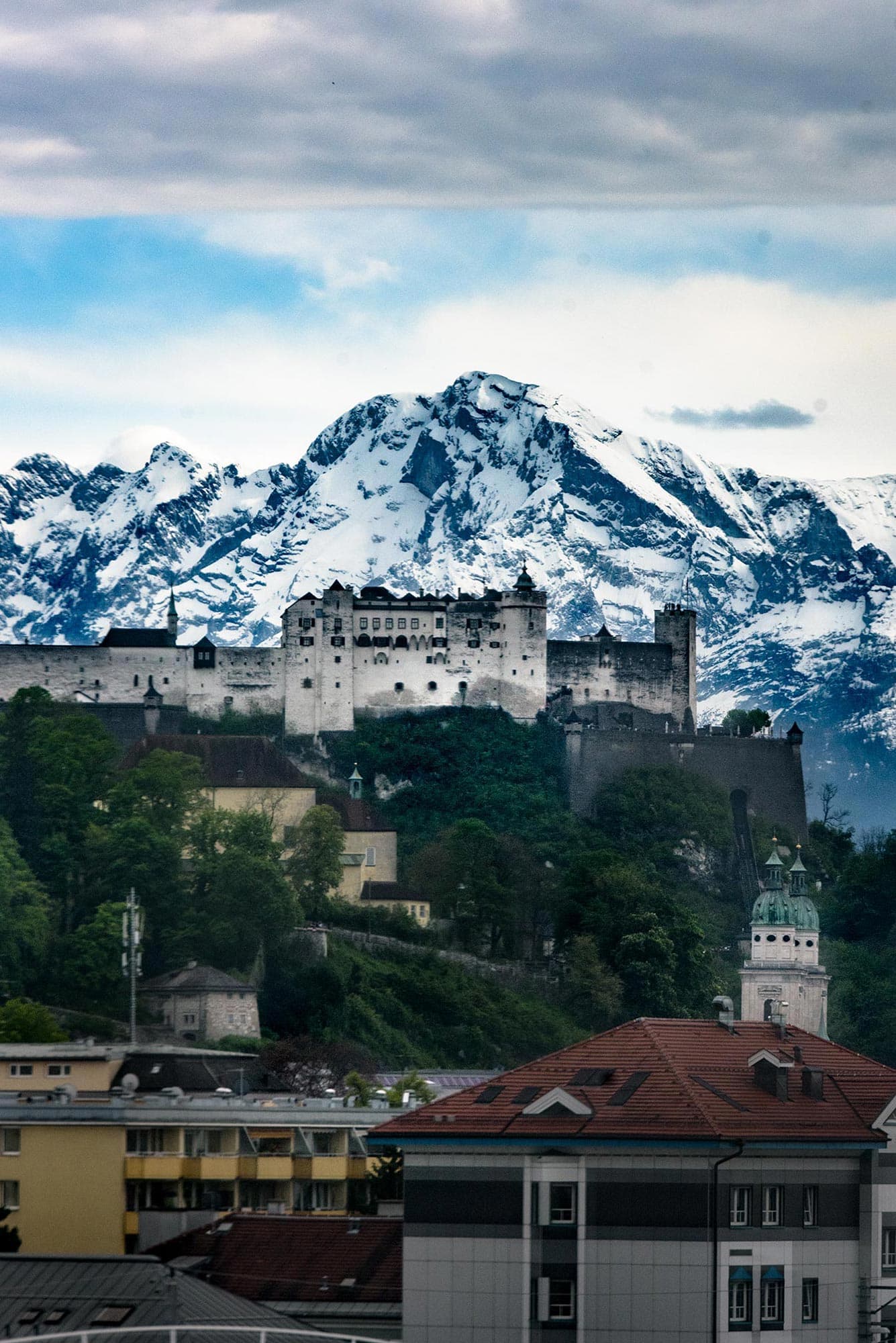 A castle sits on top of a mountain with snow capped mountains in the background.