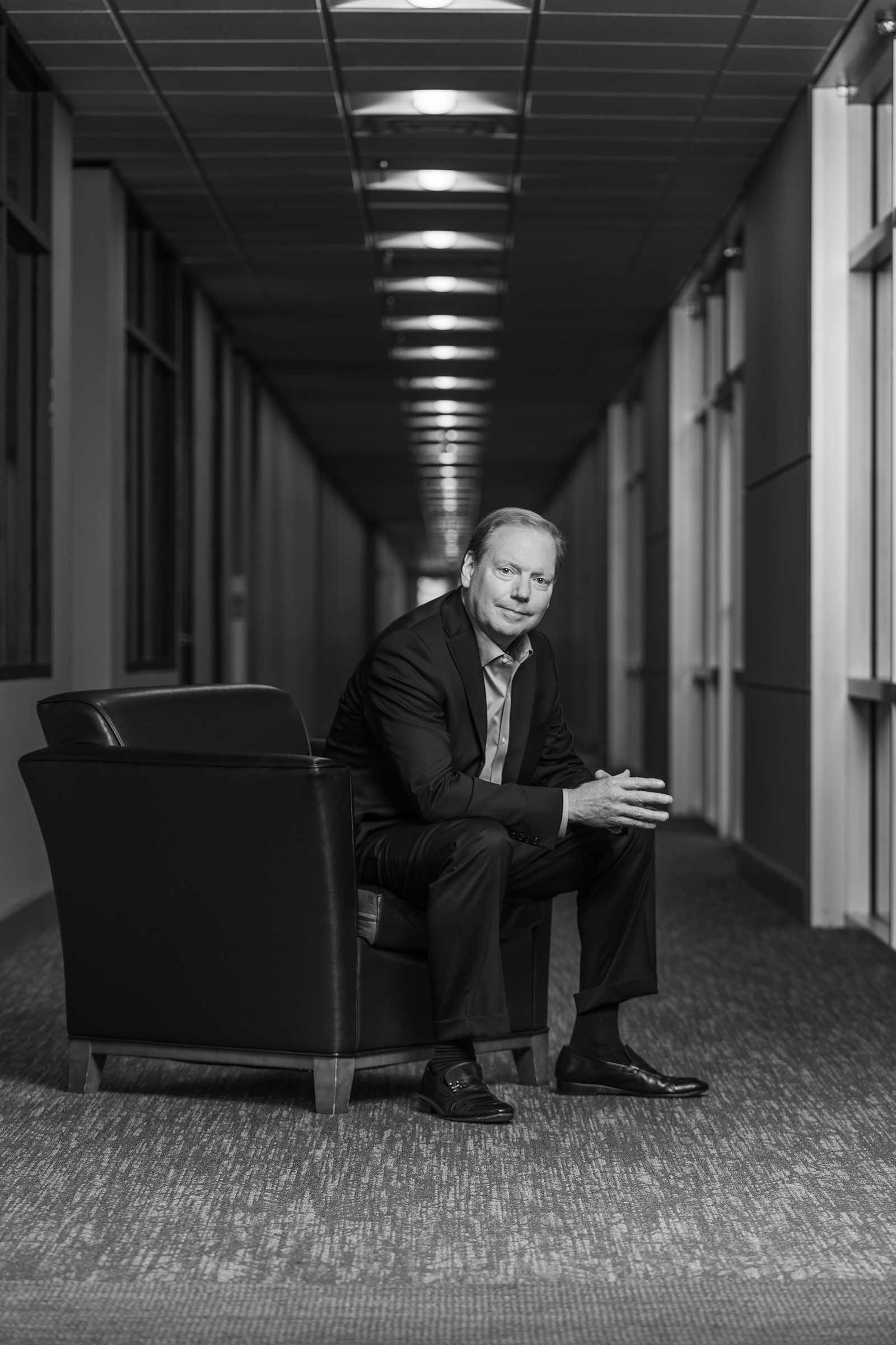 CEO poses, hand clasped in chair in hallway