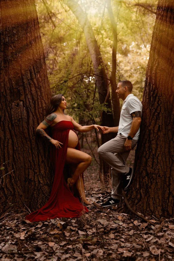 Houston Photographer captures a pregnant couple in a red dress passionately holding hands amidst the enchanting woods.